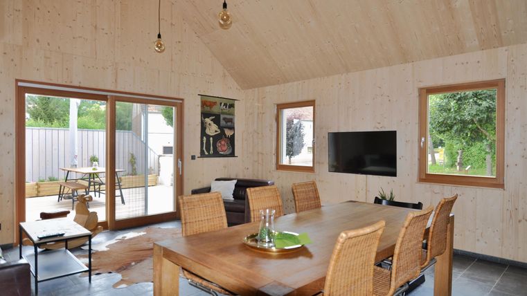 A modern living room with a large dining table and bright wood details. Large windows provide a view of the outdoors.