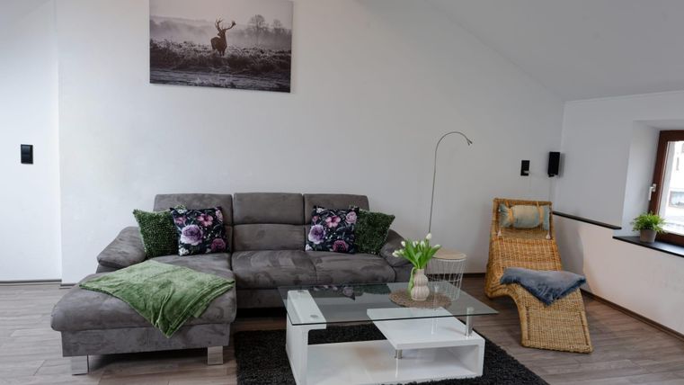 Ein gemütliches Wohnzimmer mit einem grauen Sofa und bunten Kissen. Ein Glastisch und ein Sessel ergänzen die moderne Einrichtung.