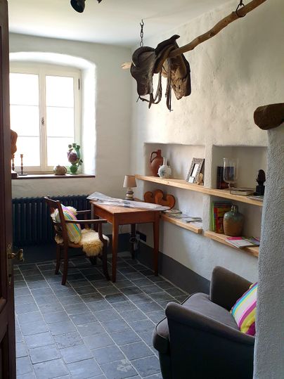 Une pièce confortable avec un bureau, un fauteuil confortable et des étagères. Sur le mur, certains chapeaux sont accrochés, et une fenêtre laisse entrer la lumière du jour.