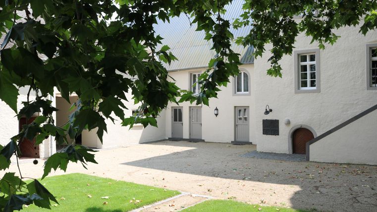 Een rustige binnenplaats met witte gebouwen en een groen gazon. Boven de binnenplaats groeien bladeren van nabijgelegen bomen.