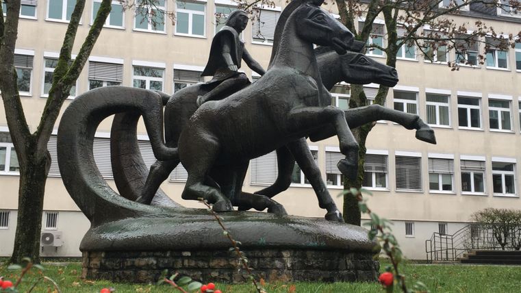 Une statue de cavalier en bronze se tient dans un espace vert devant un bâtiment. La statue montre le cavalier dans une posture calme avec une épée levée.