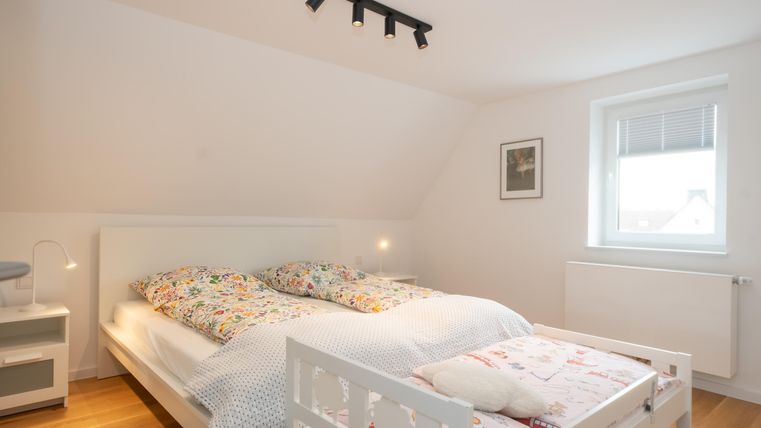 Een heldere slaapkamer met een groot bed en een extra kinderbed. De muren zijn licht, en een raam laat veel daglicht binnen.