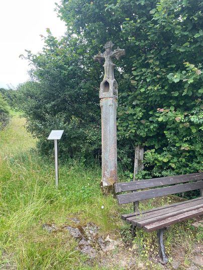 Ein altes steinernes Wegekreuz neben einer Holzbank und einem Schild, umgeben von grünen Büschen und Gras.