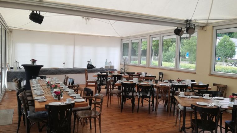 Ein eleganter Veranstaltungsraum mit Holztischen und Stühlen. Das Licht fällt durch große Fenster, und der Raum ist bereit für eine Veranstaltung.