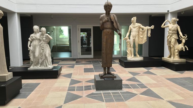 Eine Innenansicht mit mehreren Skulpturen auf Sockeln. Der Raum hat einen modernen Boden und große Fenster, die Licht hereinlassen.