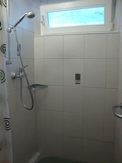 Eine helle Dusche mit weißen Fliesen und einem Fenster. Die Dusche enthält eine Handbrause und einen Duschvorhang.
