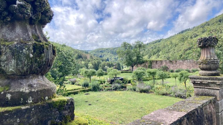 Een prachtige tuinlandschap met groene weiden en bomen. Op de achtergrond zijn zachte heuvels en een heldere lucht te zien.