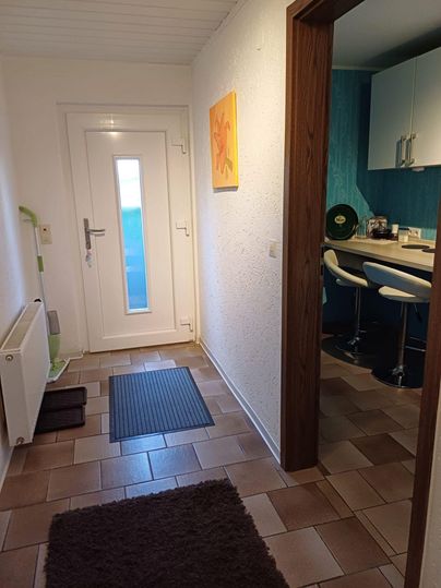 Ein heller Flur mit einer modernen Eingangstür und einem Teppich. Im Hintergrund ist eine offene Küche mit barierten Sitzplätzen zu sehen.