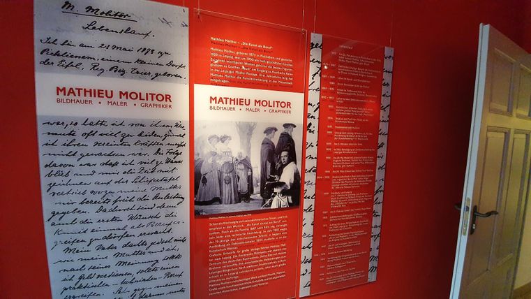 Eine Wand mit Informationen über Mathieu Molitor, einschließlich handschriftlicher Texte und Fotos. Die Wand ist rot gestrichen und zeigt verschiedene Ausstellungsstücke.