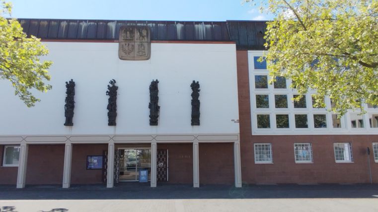 Een modern gebouw met een strakke witte gevel en een overdekte entree. Aan de gevel bevinden zich verschillende beeldhouwwerken en een wapen.