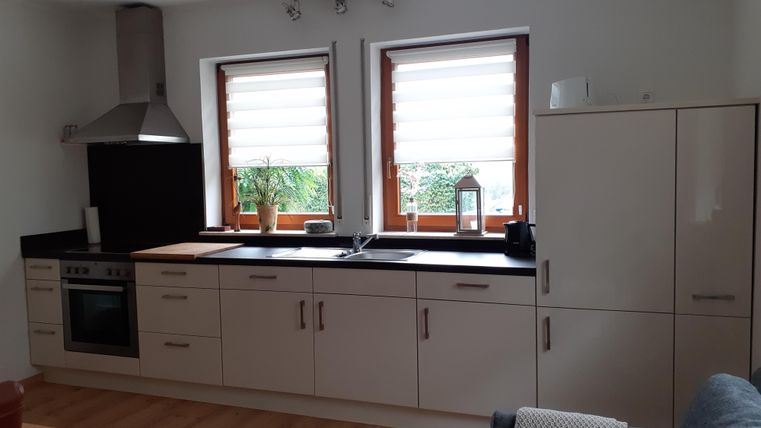 Een moderne keuken met lichte meubels en een houten werkblad. Twee ramen laten veel daglicht binnen en een plant dient als decoratie.