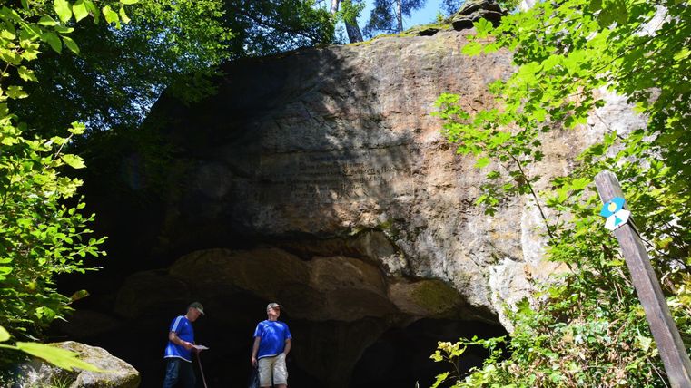 Eine natürliche Felsformation im Wald mit zwei Personen davor. Umgeben von üppigem Grün und Bäumen.