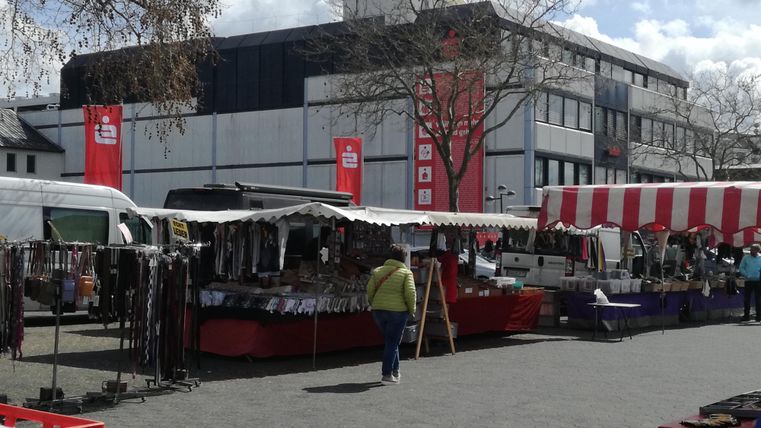 A market with various stalls and sales areas. In the background, buildings and trees are visible.
