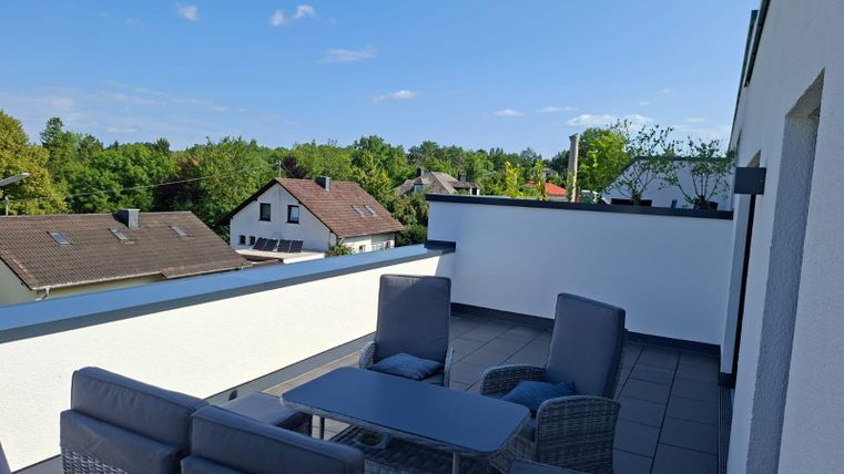 Une terrasse moderne avec des sièges et une vue sur des arbres verts et des maisons. Le ciel est clair et ensoleillé.