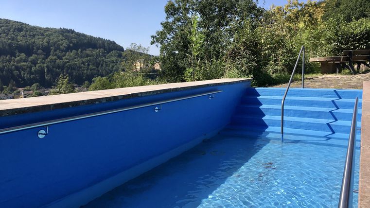 Ein blauer Pool mit einer Treppe, umgeben von Bäumen und Hügeln. Der Himmel ist klar und sonnig.