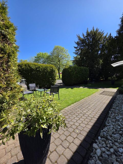 A beautiful garden with green bushes and a bright terrace. Chairs are outside under a blue sky.