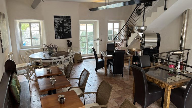 Ein gemütliches Café mit Tischen und Stühlen. Helle Fenster und eine offene Treppe sorgen für einladende Atmosphäre.