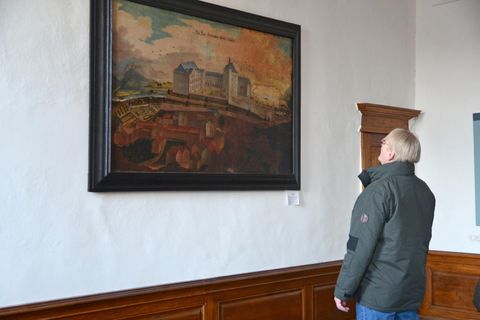 A person is looking at a painting on the wall. The image depicts an impressive landscape with historical buildings.