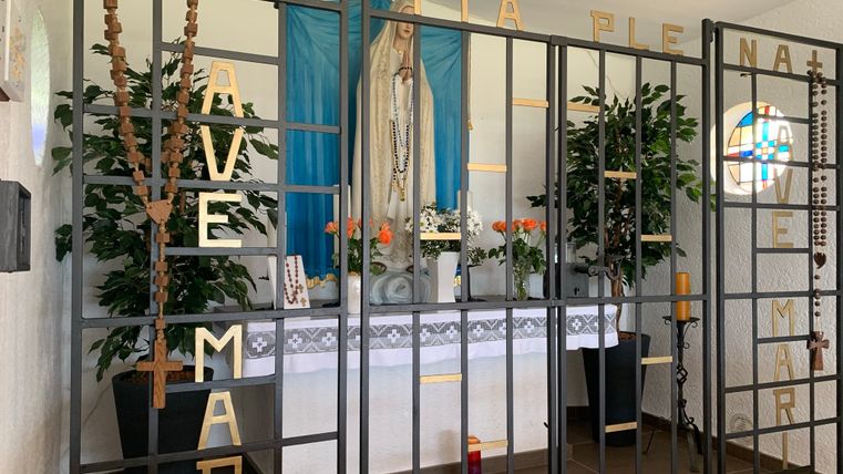 Interior view of a chapel with a statue of the Virgin Mary behind a grille with the inscription 'AVE MARIA'.