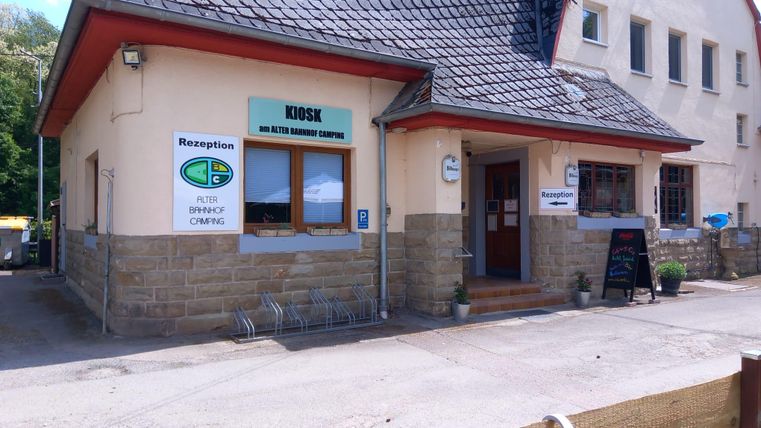 Ein Empfangsgebäude mit einem Kiosk an einem Campingplatz. Die Fassade ist einfach und einladend, umgeben von einem Parkplatz.