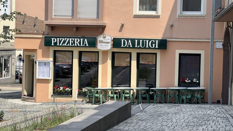 Eine Pizzeria mit dem Namen "Da Luigi" in einem hellen Gebäude. Draußen stehen grüne Stühle und Tische in einer ruhigen Umgebung.