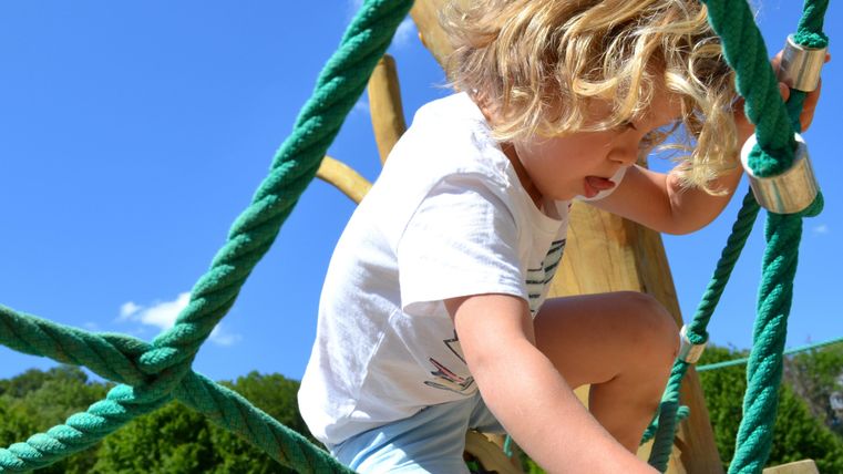 Ein Kind spielt auf einem Klettergerüst aus Seilen. Die Sonne scheint und der Himmel ist blau.