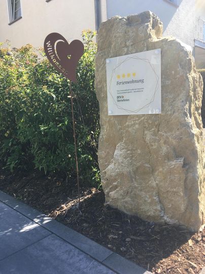 Een grote steen met een welkomstbord en een hartsymbool staat in een tuinlandschap. De omgeving is groen en vriendelijk ingericht.