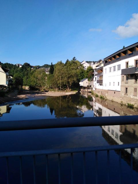 Ein ruhiger Fluss mit Spiegelungen der Gebäude und Bäume am Ufer. Der Himmel ist klar und blau, was eine friedliche Atmosphäre schafft.