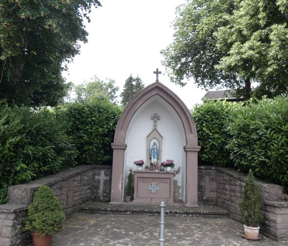 Small chapel with a statue of the Virgin Mary, surrounded by trees and bushes. The chapel has a cross on the roof and flowers inside., © TI Bitburger Land