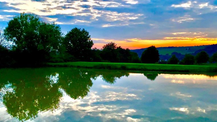 Ein ruhiger See mit spiegelndem Wasser und umgebenden Bäumen. Der Himmel leuchtet in warmen Farben bei Sonnenuntergang.