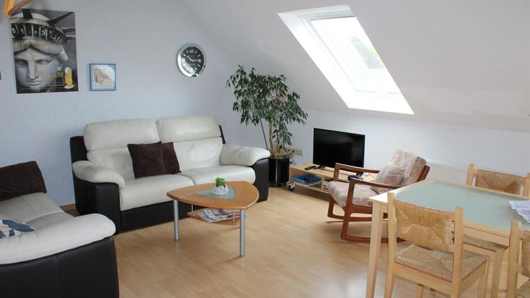 Ein gemütliches Wohnzimmer mit einer hellen Einrichtung. Es gibt ein Sofa, einen Esstisch und viel Tageslicht durch ein Dachfenster.