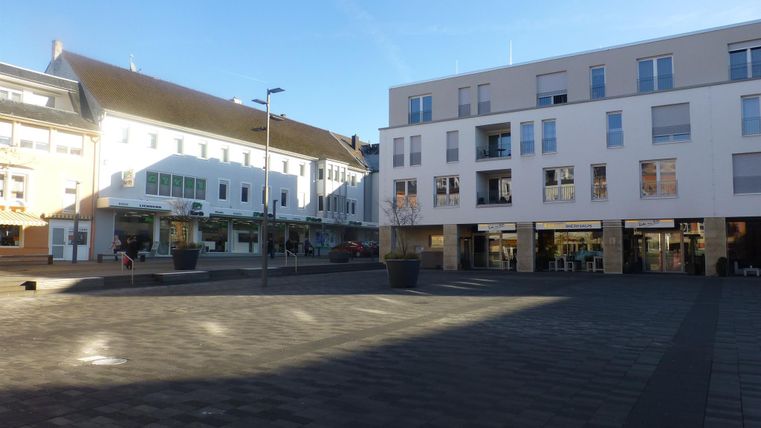 Ein heller Platz mit modernen Gebäuden und Geschäften. Der Himmel ist blau und die Umgebung wirkt einladend.