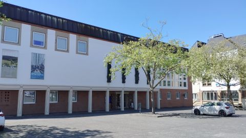 A modern building with large windows and a bright, clear sky. In front of the building, there are some trees and parked cars.