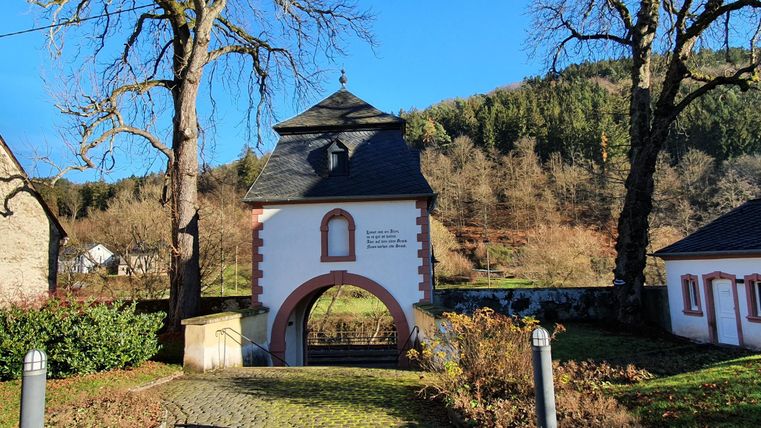Een historisch poortgebouw met een boog, omringd door bomen en zachte heuvels. De lucht is helder en blauw.