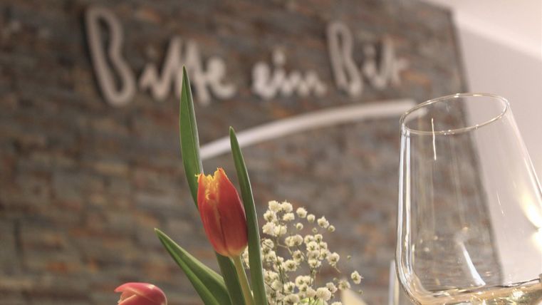 Ein Glas Wein steht neben einem Blumenarrangement mit frischen Tulpen. Im Hintergrund ist eine elegante Wand mit der Aufschrift "Bitte ein Bit" zu sehen.