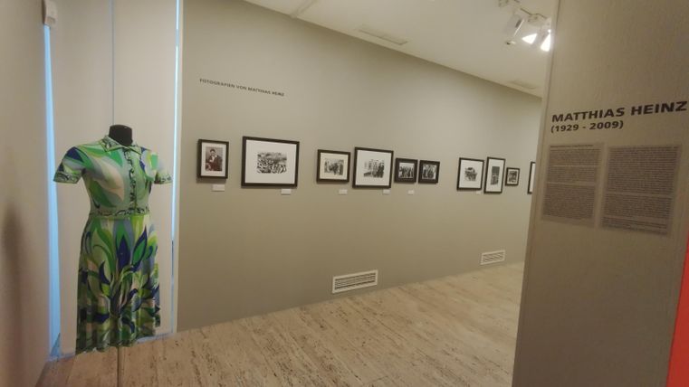 An exhibition space with a green dress on a stand and several pictures on the wall. The exhibition showcases works by Matthias Heinz.