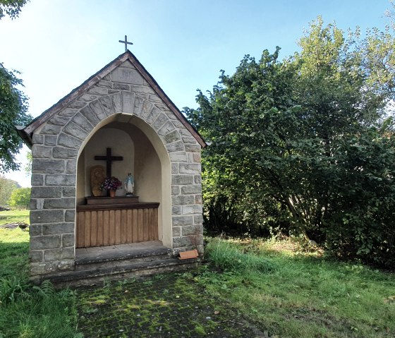 Een kleine stenen kapel met een kruis en een beeld van de Maagd Maria, omringd door groene bomen en gras., © TI Bitburger Land