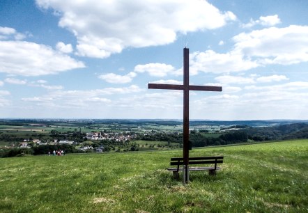 Croix en bois sur une prairie avec un banc, en arrière-plan un vaste paysage et un village sous un ciel bleu avec des nuages., © TI Bitburger Land