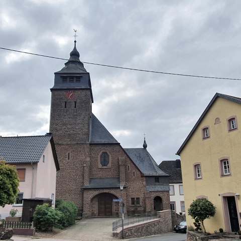 Dorfplatz mit einer Kirche aus Backstein, umgeben von bunten Häusern. Ein Baum im Vordergrund, bewölkter Himmel im Hintergrund., © TI Bitburger Land S.Wagner