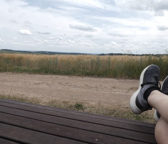 Person liegt auf einer Bank, die Beine ausgestreckt. Im Hintergrund ein Feld und bewölkter Himmel., © TI Bitburger Land