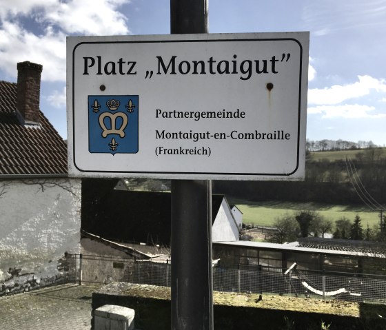 Sign 'Platz Montaigut' with coat of arms, reference to partner municipality Montaigut-en-Combraille. Rural landscape in the background., © TI Bitburger Land