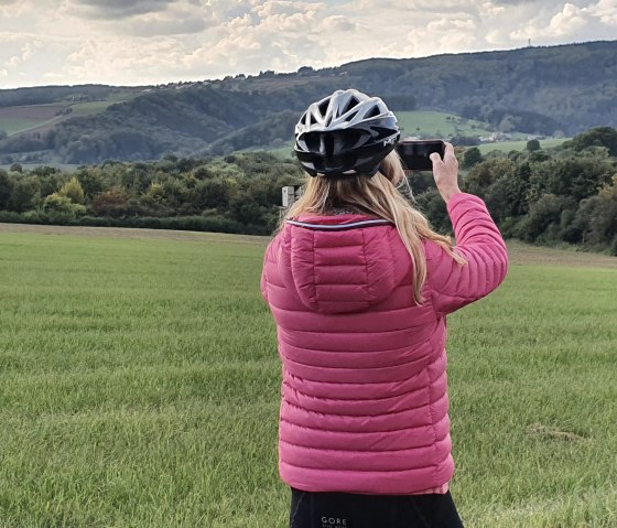 Een persoon met een roze jas en fietshelm fotografeert een groen landschap met heuvels op de achtergrond., © TI Bitburger Land