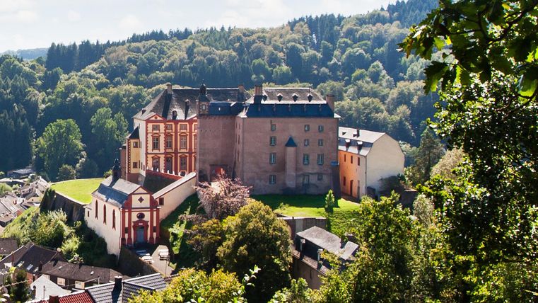 Eine beeindruckende Burg umgeben von grünen Wäldern und sanften Hügeln. Klarer blauer Himmel und idyllische Landschaft verleiht der Szene eine ruhige Atmosphäre.