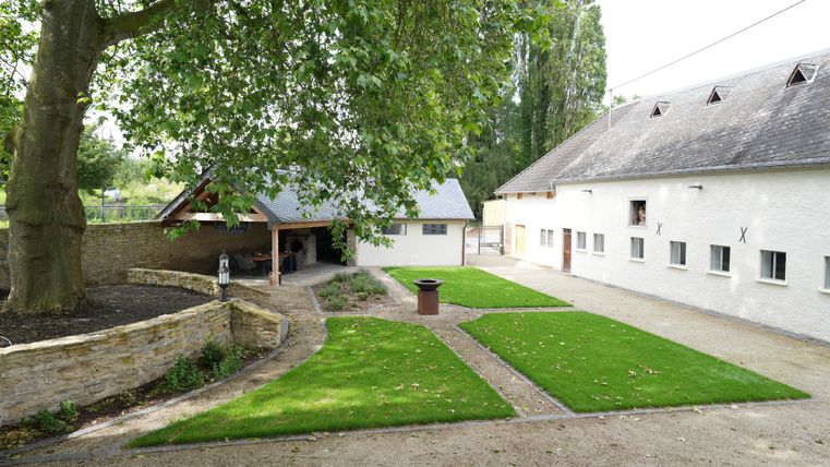 Een rustige binnenplaats met groen gras, bomen en een bijgebouw. Op de achtergrond zijn oude stallen te zien.