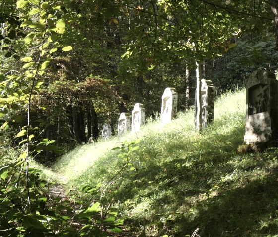 Een schaduwrijk bospad met stenen kruiswegstaties op de helling. Zonlicht valt door de bomen op het gras., © TI Bitburger Land