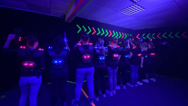 Eine Gruppe von Personen steht in einem dunklen Raum mit leuchtenden Lasern. Sie trägt spezielle Westen und bereitet sich auf ein Laserspiel vor.