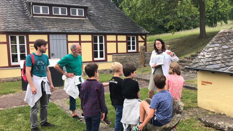 Een groep kinderen luistert naar een vrouw die iets uitlegt. Op de achtergrond is een historisch gebouw in een groene omgeving te zien.