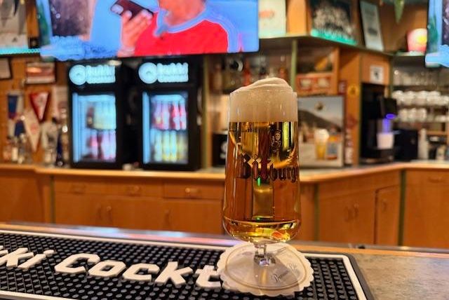 Ein Glas Bier steht auf der Theke in einer Bar. Im Hintergrund sind Getränkeautomaten und Fernseher zu sehen.