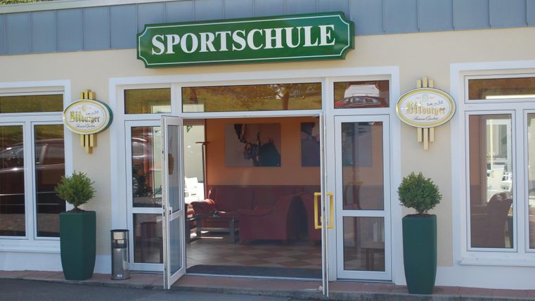 Ein einladender Eingang zu einer Sportschule mit großen Fenstern. Die Fassade ist hell und modern gestaltet.