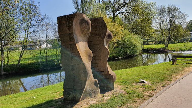Steinskulptur am Flussufer mit Bäumen im Hintergrund.